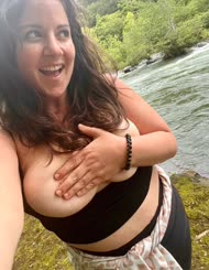 A woman with big breasts takes a selfie by a river with the water looking muddy