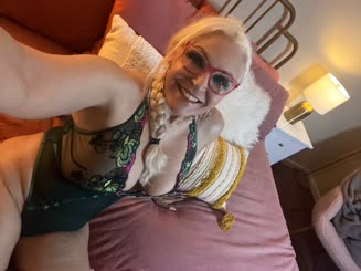 A woman with a braid in her hair takes a selfie in a bed wearing a short dress and glasses
