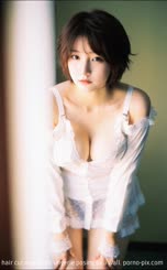 A woman with a short hair cut and white lingerie posing by a wall.