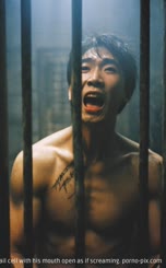 There is a shirtless man with a tattooed neck standing in front of a jail cell with his mouth open as if screaming.