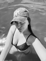 A woman in a bathing suit with her hair in pig tails takes a selfie in the water wearing a Yankees cap