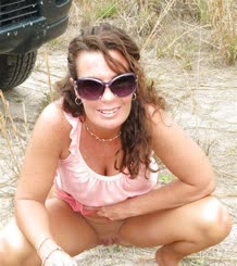 A woman wearing a pink shirt and sunglasses squatting on the beach.